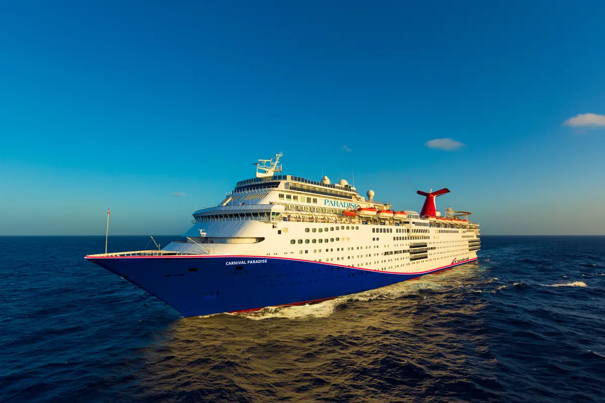 Die Carnival Paradise, das große weiß-blaue Schiff mit rotem Schornstein, fährt auf blauem Meer unter klarem Himmel.