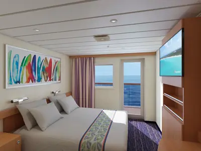 Innenansicht einer modernen Kabine auf der Carnival Paradise mit einem Doppelbett. Ein großes, farbenfrohes Kunstwerk mit Surfbrettern hängt über dem Bett. Rechts sind ein Fenster und eine Tür, die zu einem Balkon mit Meerblick fuehren. Ein großer Fernseher ist an der Wand befestigt.