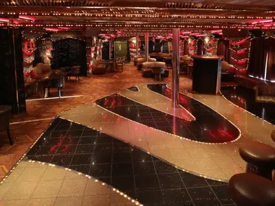 Ansicht eines luxuriösen Nachtclubs auf der Carnival Paradise. Eine glänzende Tanzfläche mit LED-Lichtstreifen in Schwarz und Gold dominiert den Raum. Eine Tanzstange steht mittig. Ringsherum sind bequeme Sessel und Sofas sowie Bartische und Hocker platziert. Rote Akzentbeleuchtung sorgt für eine lebhafte Atmosphäre.