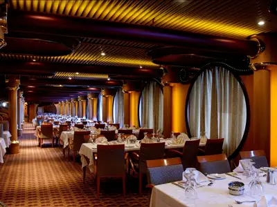 Innenansicht eines langen und eleganten Restaurants auf der Carnival Paradise. Mehrere Reihen gedeckter Tische mit Stühlen, Holzsäulen und goldgelb beleuchteten Wänden mit runden Fenstern.