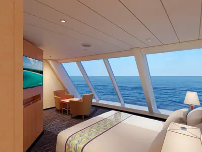 Geräumige Suite der Carnival Paradise mit schräg angeordneten Panoramafenstern mit direktem Meerblick. Ein Doppelbett, ein kleiner Tisch mit zwei Sesseln und ein Fernseher sind sichtbar.