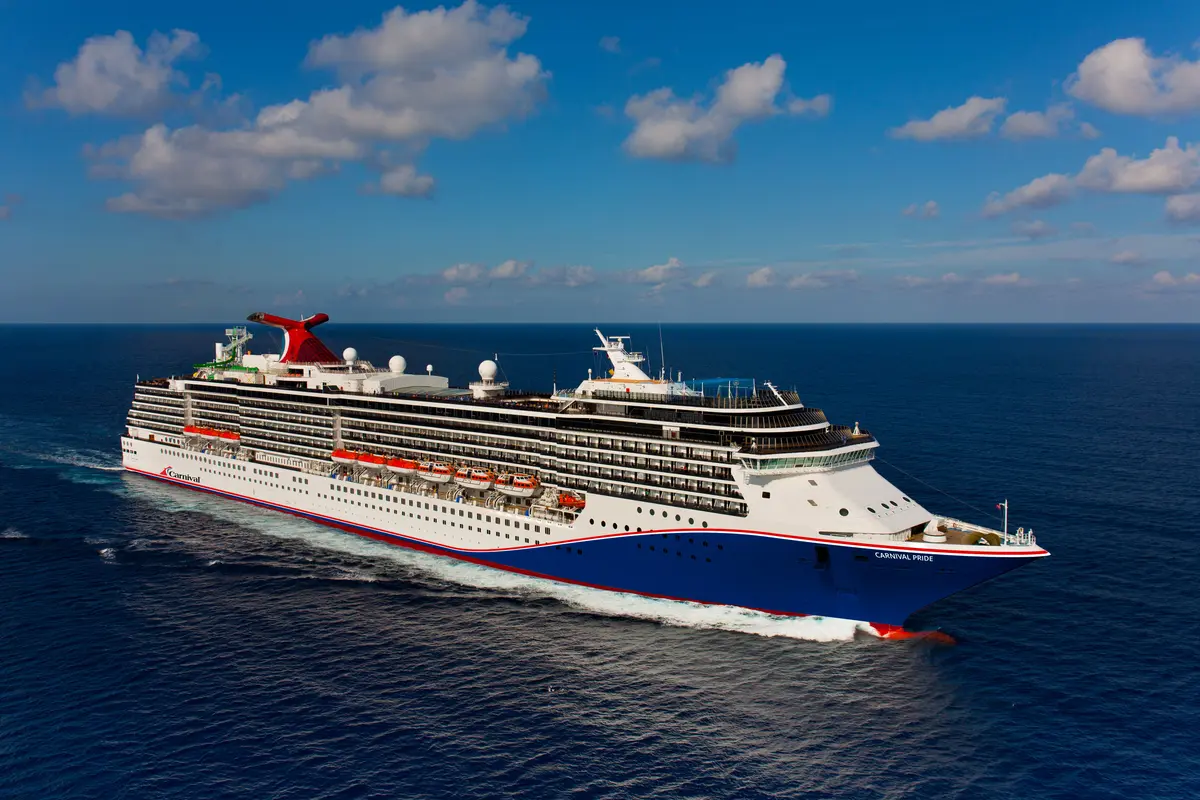 Luftaufnahme der Carnival Pride von der Seite, auf dem offenen Meer fahrend. Das Schiff hat einen roten Schornstein, eine blaue Bugpartie und ist umgeben von blauem Wasser unter einem blauen Himmel mit weißen Wolken.