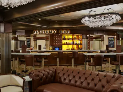 Innenansicht der Alchemy Bar auf der Carnival Pride. Die Theke ist umgeben von braunen Barhockern. Im Vordergrund ein braunes Chesterfield-Sofa. Eine ovale Kristall-Lichtinstallation hängt von der Decke.
