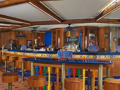 Die Blue Iguana Tequila Bar an Bord der Carnival Pride. Die Bar hat eine holzverkleidete Wand, eine blaue Theke mit gelb-blauen Fliesen und bunte Barhocker. Flaschen und Zapfanlagen sind sichtbar.