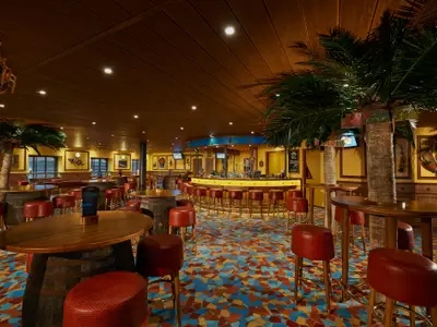 Innenansicht einer thematischen Bar auf der Carnival Pride mit Palmen, Fass-Tischen, roten Hockern und einem lebhaften Mosaikboden. Eine kreisrunde Bar ist im Hintergrund zu sehen. Die Beleuchtung schafft eine warme, tropische Atmosphäre und lädt zum Verweilen ein.