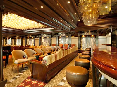 Elegante Bar-Lounge auf der Carnival Pride. Glänzender Holz-Bartresen mit Hockern rechts, bequeme Sessel und lange Sitzbank links. Opulenter Kronleuchter und dunkles Holzinterieur. Ohne Personen.