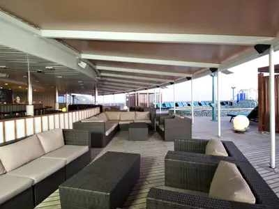 Überdachte Outdoor-Lounge auf der Carnival Pride. Dunkelbraune Rattansofas, helle Kissen. Im Hintergrund Bar mit blauen Akzenten und Poolbereich mit Liegen.