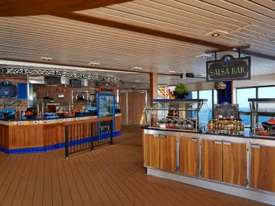 Ansicht des mexikanischen Buffet-Restaurants 'Blue Iguana Cantina' an Bord der Carnival Pride. Es gibt eine Theke mit Tacos und eine Salsa Bar mit verschiedenen Soßen und Beilagen. Im Hintergrund ist das Meer sichtbar. Der Raum ist hell beleuchtet und modern eingerichtet, mit Holz- und Fliesenakzenten.