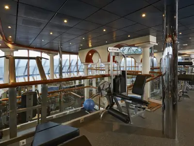 Modernes Fitnessstudio auf der Carnival Pride mit Kraftmaschinen, Hanteln und blauem Gymnastikball. Große Fenster bieten weite Sicht auf das Meer. Helle Raumgestaltung mit Säulen und Holzelementen.