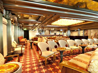 Die elegante Schiffs-Lounge zeigt bequeme Stühle und Sofas, kleine Tische, einen Kronleuchter und eine Bar im Hintergrund. An den Wänden sind Gemälde und Säulen zu sehen.
