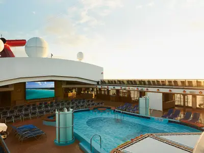 Blick auf das obere Deck der Carnival Pride bei Sonnenuntergang. Ein großer Pool mit Barhockern im Wasser, umgeben von Liegestühlen und Sitzreihen. Im Hintergrund eine riesige LED-Leinwand, die einen tropischen Strand zeigt. Darüber die Schiffsaufbauten mit Radomen.