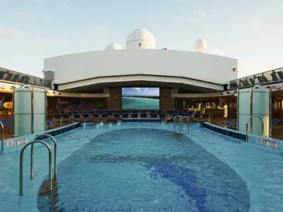 Weitwinkelaufnahme des menschenleeren Pooldecks der Carnival Pride am Tag. Ein großer Bildschirm zeigt eine tropische Strandlandschaft. Zahlreiche Liegestühle umgeben den Pool, dessen Wasser leicht gewellt ist. Im Hintergrund sind Schiffstrukturen und der blaue Himmel zu sehen.