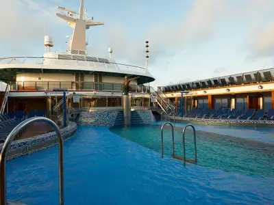 Ein weitläufiger Poolbereich an Bord der Carnival Pride mit blauen Liegestühlen, Schwimmbecken und einer bronzenen Seemanns-Skulptur. Das Meer ist im Hintergrund sichtbar.