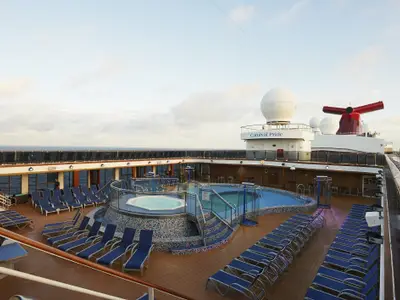 Sonnendeck der Carnival Pride mit Pool, Whirlpool und Liegestühlen. Im Hintergrund das Meer und der obere Schiffsaufbau mit Radomen und rotem Schornstein.