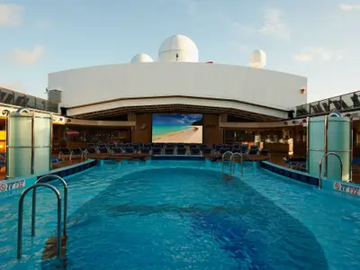 Blick auf den Hauptpoolbereich der Carnival Pride. Ein großer, beleuchteter LED-Bildschirm zeigt eine tropische Strandlandschaft. Mehrere Reihen blauer Liegestühle sind vor dem Bildschirm auf einer erhöhten Plattform platziert. Das Schwimmbecken mit blauem Wasser erstreckt sich im Vordergrund, flankiert von Poolleitern und einer Duschsäule. Die obere Decks sind mit weißen Aufbauten und Antennenkugeln sichtbar.