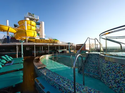 Weitwinkelaufnahme eines sonnigen Pooldecks der Carnival Pride mit Pool, Hot Tub, gelben Wasserrutschen und türkisfarbenen Liegestühlen unter blauem Himmel. Meer im Hintergrund.