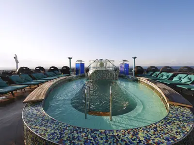 Ovaler Poolbereich auf der Carnival Pride mit türkis gepolsterten Liegestühlen und Meerblick unter blauem Himmel. Ein Whirlpool befindet sich am Ende des Pools.