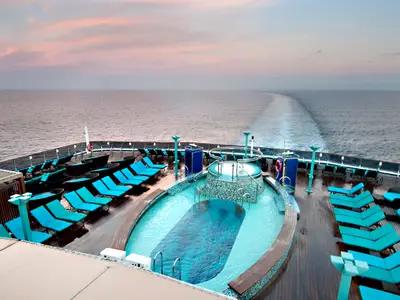 Blick auf das Heckdeck der Carnival Pride im Sonnenuntergang. Pool, Whirlpool, türkisblaue Liegestühle auf Holzdeck. Weites Meer mit Kielwelle und rosafarbenem Himmel.