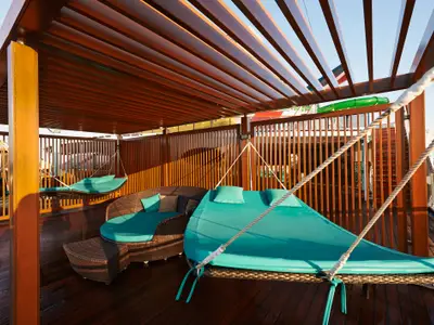 Drei türkisfarbene Hängematten-Liegen unter einer Holzpergola auf einem Deck der Carnival Pride mit Meerblick. Braune Holzvertäfelung und dunkler Holzboden.