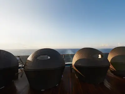 Vier moderne, geflochtene Lounge-Sessel in dunklem Ton stehen auf einem hölzernen Sonnendeck mit Reling und Blick auf das weite, blaue Meer und den Horizont, unter einem hellen Himmel.