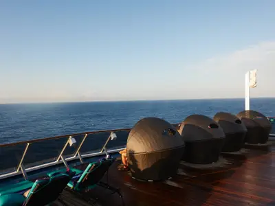 Sonnendeck der Carnival Pride mit dunklen Lounge-Sesseln und einer Person, die auf das blaue Meer blickt. Holzdeck und Glasgeländer unter klarem Himmel.