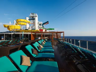 Blick auf das Sonnendeck der Carnival Pride mit gelben Wasserrutschen und türkisfarbenen Liegestühlen. Im Hintergrund das weite Meer unter blauem Himmel. Einige Personen entspannen an Bord.