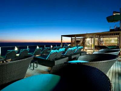 Abendstimmung auf dem Sonnendeck der Carnival Pride. Mehrere Lounge-Liegen mit türkisen Auflagen stehen auf Holzplanken. Im Hintergrund ist das Meer und ein farbenfroher Himmel beim Sonnenuntergang zu sehen. Rechts oben eine Wasserrutsche.