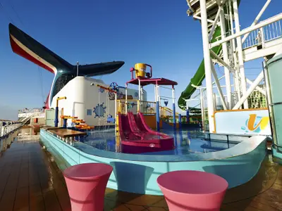 Blick auf das bunte Carnival WaterWorks mit pinken Rutschen, Wasserspielen und dem charakteristischen roten Schornstein auf dem Oberdeck der Carnival Pride bei Tageslicht.