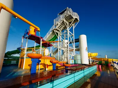 Ein lebhaftes Wasserspielparadies an Bord der Carnival Pride mit mehreren bunten Wasserrutschen, Eimern und Spritzanlagen unter strahlend blauem Himmel. Ohne Personen.