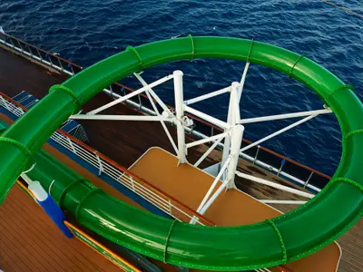 Eine grüne, geschwungene Wasserrutsche der Carnival Pride, die sich über einem dunklen, welligen Meer befindet. Der obere Teil des Schiffsdecks mit Holzbelag und weißen Geländern ist ebenfalls sichtbar.