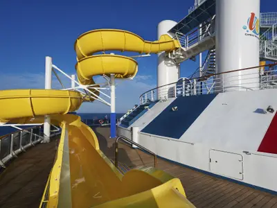 Sonnige Ansicht von gelben Wasserrutschen auf dem Deck der Carnival Pride. Hintergrund: blaues Meer und Himmel. Weiße Stützen, rot-weiß-blaue Schiffselemente sichtbar.