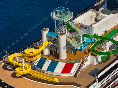 Luftaufnahme der Carnival Pride mit mehreren gelben und grünen Wasserrutschen auf dem oberen Deck. Im Hintergrund ist das blaue Meer zu sehen. Einige Sonnenliegen sind platziert und vereinzelt sind Personen zu erkennen, die sich sonnen oder umherlaufen.