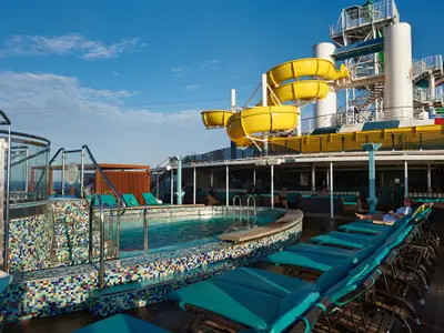 Oberes Deck der Carnival Pride mit Pool, bunten Mosaikfliesen und türkisen Liegestühlen. Leuchtend gelbe Wasserrutschen im Hintergrund. Eine Person entspannt auf einer Liege.