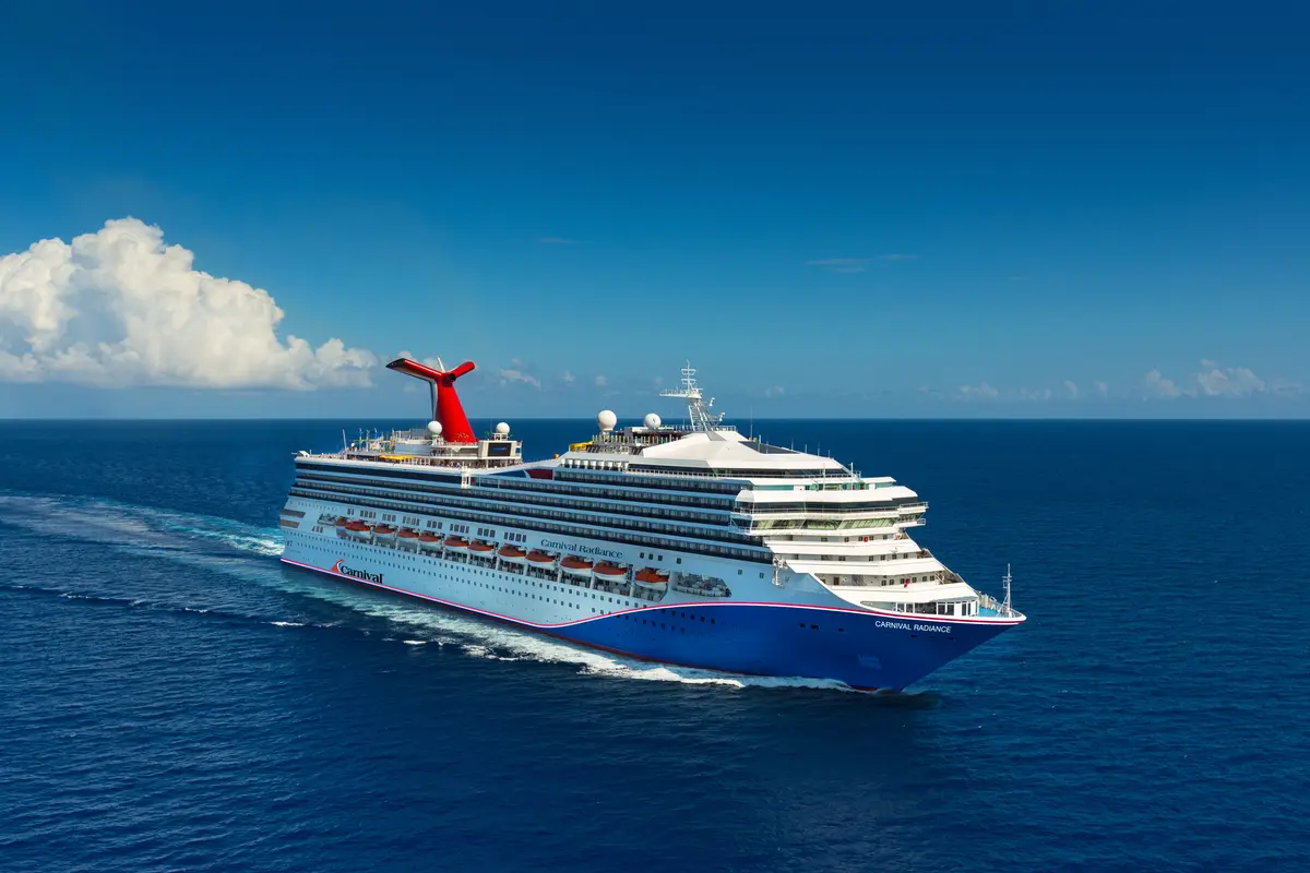 Die Carnival Radiance gleitet unter blauem Himmel mit einigen weißen Wolken über das offene Meer, eine Bugwelle hinter sich lassend. Die markanten roten Schornsteine und die blau-weiße Bemalung sind gut zu erkennen.