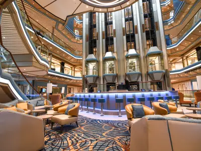 Ein modernes Atrium der Carnival Radiance mit einer leuchtenden Bar, komfortablen Lounge-Sesseln in Blau und Orange, und gläsernen Aufzügen, die sich über mehrere Decks erstrecken.