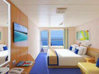 Eine helle, moderne Balkonkabine an Bord der Carnival Radiance mit einem Doppelbett, Sitzecke und Fernseher. Durch die Glastür und das Fenster ist das weite blaue Meer und der Himmel sichtbar.