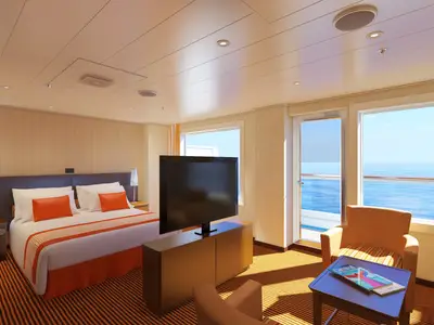 Helle, moderne Balkonkabine auf der Carnival Radiance mit Doppelbett, orangefarbenen Akzentkissen, TV und Sitzbereich mit Meerblick durch die Fenstertür.
