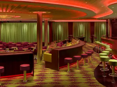 Weitwinkelansicht einer modernen Bar und Lounge auf der Carnival Radiance. Der Raum hat eine geschwungene Bar mit neonroter Beleuchtung, roten und lindgrünen Hockern. Im Hintergrund gibt es eine Bühne mit grünen Vorhängen und rote Theatersitze.