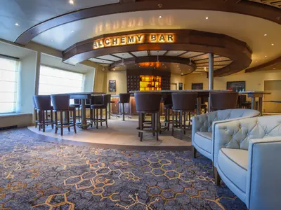 Eine einladende Alchemy Bar auf der Carnival Radiance mit einer geschwungenen Theke, hohen Hockern, kleinen Tischen und bequemen, hellen Sesseln. Große Fenster und warmes, indirektes Licht schaffen eine gemütliche Atmosphäre. Der Teppich hat ein geometrisches Muster.