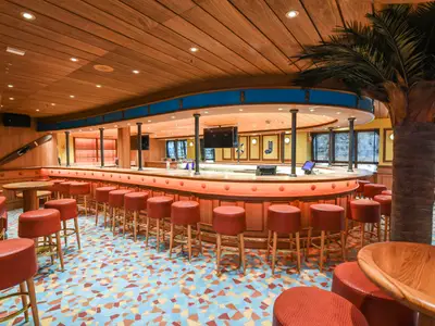 Eine leere Bar-Lounge im maritimen Stil mit langer Theke, roten Barhockern, Holzwänden und einer Palme. Der Boden hat ein buntes Mosaikmuster.