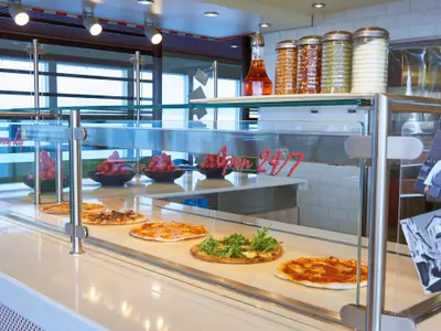 Eine Pizzastation an Bord der Carnival Radiance zeigt frische Pizzen und Schüsseln mit Tomaten und Rucola unter einer Glasvitrine. Im Hintergrund ein Fenster mit Meerblick.