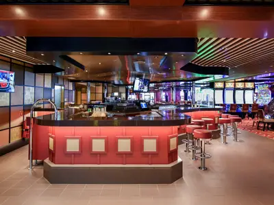 Blick in ein modernes Casino der Carnival Radiance mit einer halbrunden Bar mit roten Hockern im Vordergrund und zahlreichen leuchtenden Spielautomaten im Hintergrund.