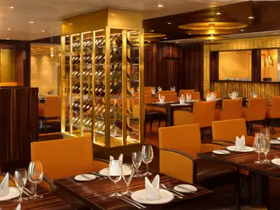 Elegantes Kreuzfahrtrestaurant mit gedeckten Holztischen, orangefarbenen Stühlen und einer goldfarbenen Weinlagerung. Eine offene Küche mit goldenen Hängelampen im Hintergrund.