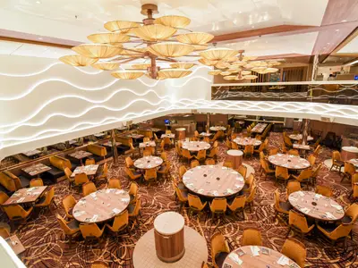 Luftaufnahme eines modernen, mehrstöckigen Restaurants auf der Carnival Radiance mit zahlreichen runden und rechteckigen Tischen, umgeben von gold-braunen Stühlen. Die Decke ist mit großen, goldenen Leuchten und wellenförmigen, beleuchteten Paneelen gestaltet.