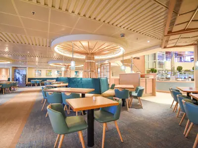 Helles, modernes Restaurant der Carnival Radiance. Holztische, teal-farbene Stühle und Bänke. Im Hintergrund Buffetbereich und Treppe. Decke mit Holzlamellen und runden Lichtinstallationen.
