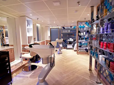Ein moderner, heller Friseursalon auf der Carnival Radiance mit zwei Haarwaschbecken, Frisierstühlen und Regalen voller Beauty-Produkte. Der Raum ist in hellen Farben mit Holzboden und blauen Akzenten gehalten.