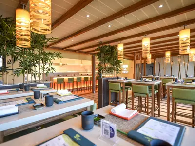 Innenansicht eines modernen japanischen Spezialitätenrestaurants auf der Carnival Radiance. Mehrere Tische mit grünen Stühlen, umgeben von Bambuspflanzen. Im Hintergrund eine beleuchtete Sushi-Bar mit Barhockern.