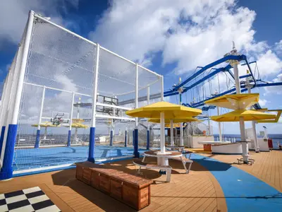 Outdoor-Sportdeck auf der Carnival Radiance mit einem umnetzten Basketballplatz, einem blauen Seilgarten und gelben Sonnenschirmen. Holzboden und blauer Himmel mit Wolken.