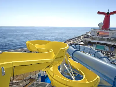 Gelbe und blaue Wasserrutschen auf dem Oberdeck der Carnival Radiance unter blauem Himmel mit Blick auf das offene Meer. Der rote Schornstein und ein Poolbereich sind sichtbar.