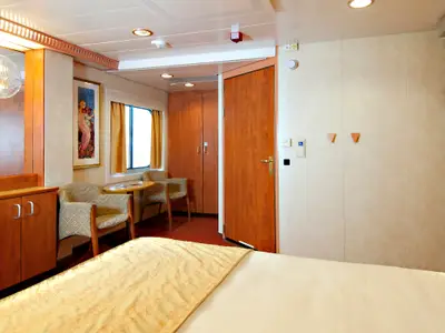 Geräumige Außenkabine auf der Carnival Spirit. Links ein Schreibtisch mit TV, Stühlen und einem Fenster. Rechts ein Doppelbett mit Nachttischlampe.